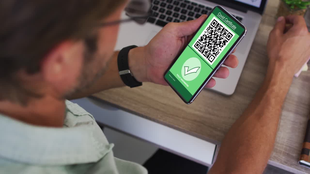Man at desk holding smartphone with covid vaccination certificate and qr code on screen