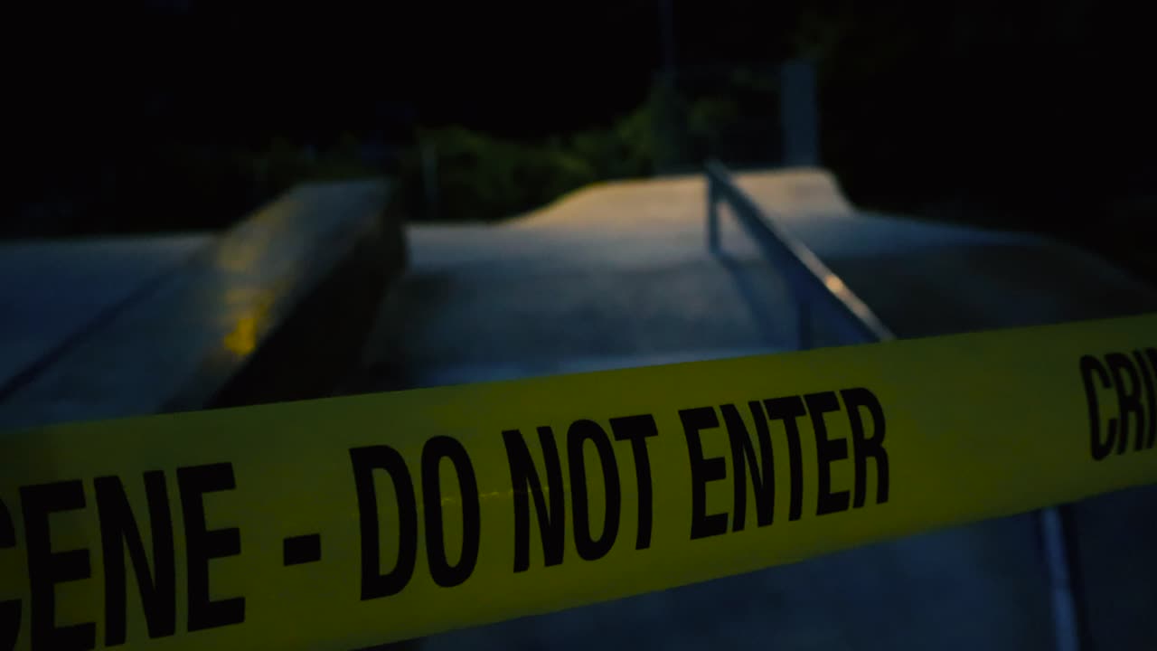 Close up footage glides slowly along fluttering yellow crime scene tape pulled across the skatepark ramps on a moody night. Somber park concrete surface and bushes behind are dimly lit by streetlight