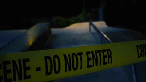 close up footage glides slowly along fluttering yellow crime scene tape pulled across the skatepark ramps on a moody night. somber park concrete surface and bushes behind are dimly lit by streetlight