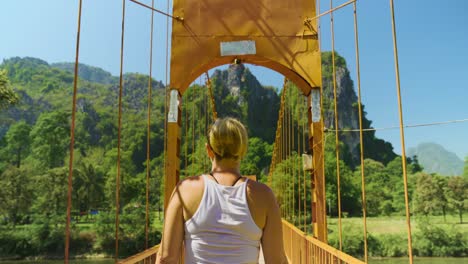 vista trasera de una mujer joven cruzando un puente peatonal colgante sobre el río nam song en vang vieng, laos