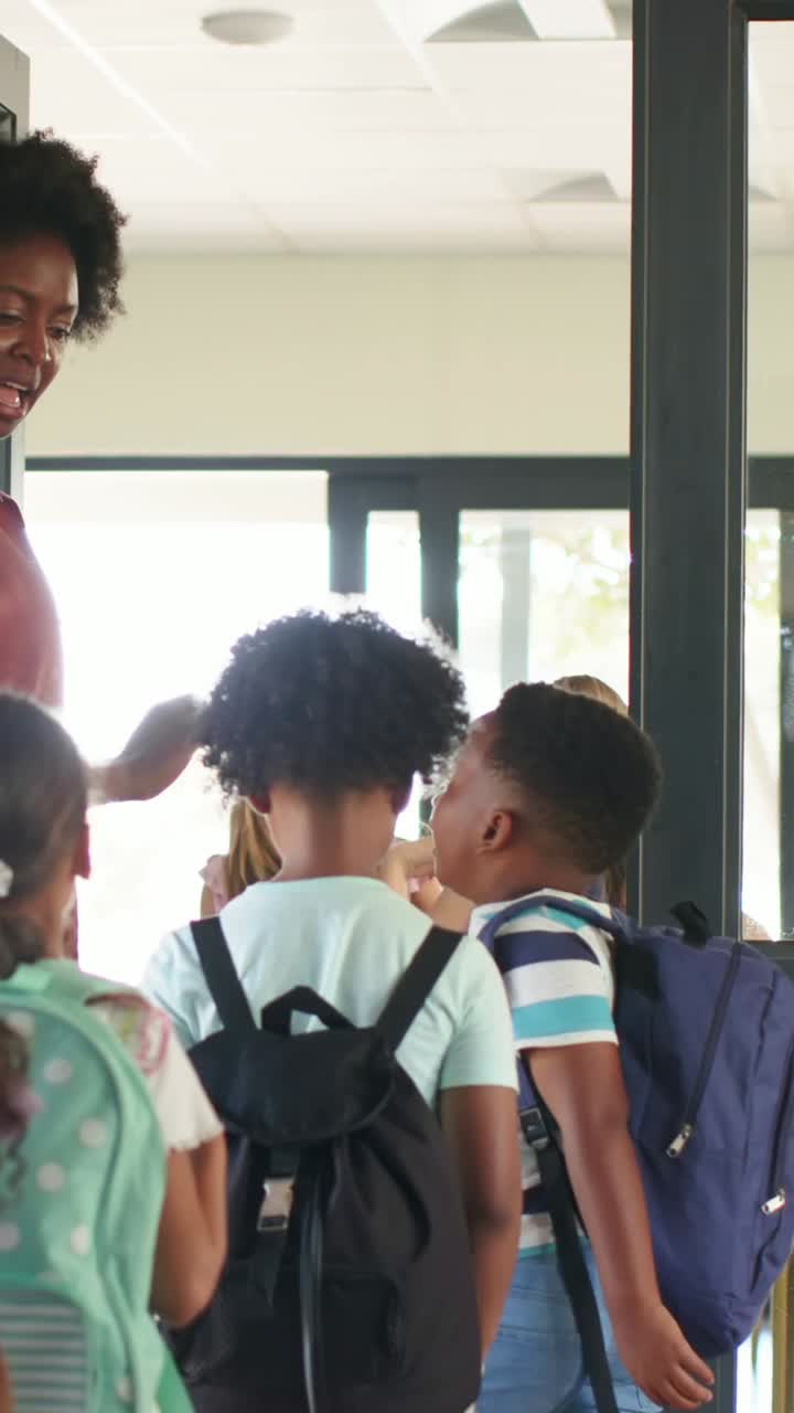 Video verticale: Insegnante con top color ruggine che saluta e dà il cinque ai bambini che entrano a scuola con gli zaini.