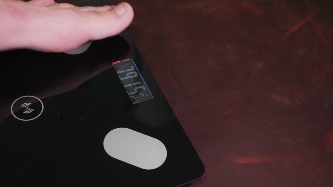 A person's feet stand on a black digital scale displaying weight on a dark brown wooden floor indoors