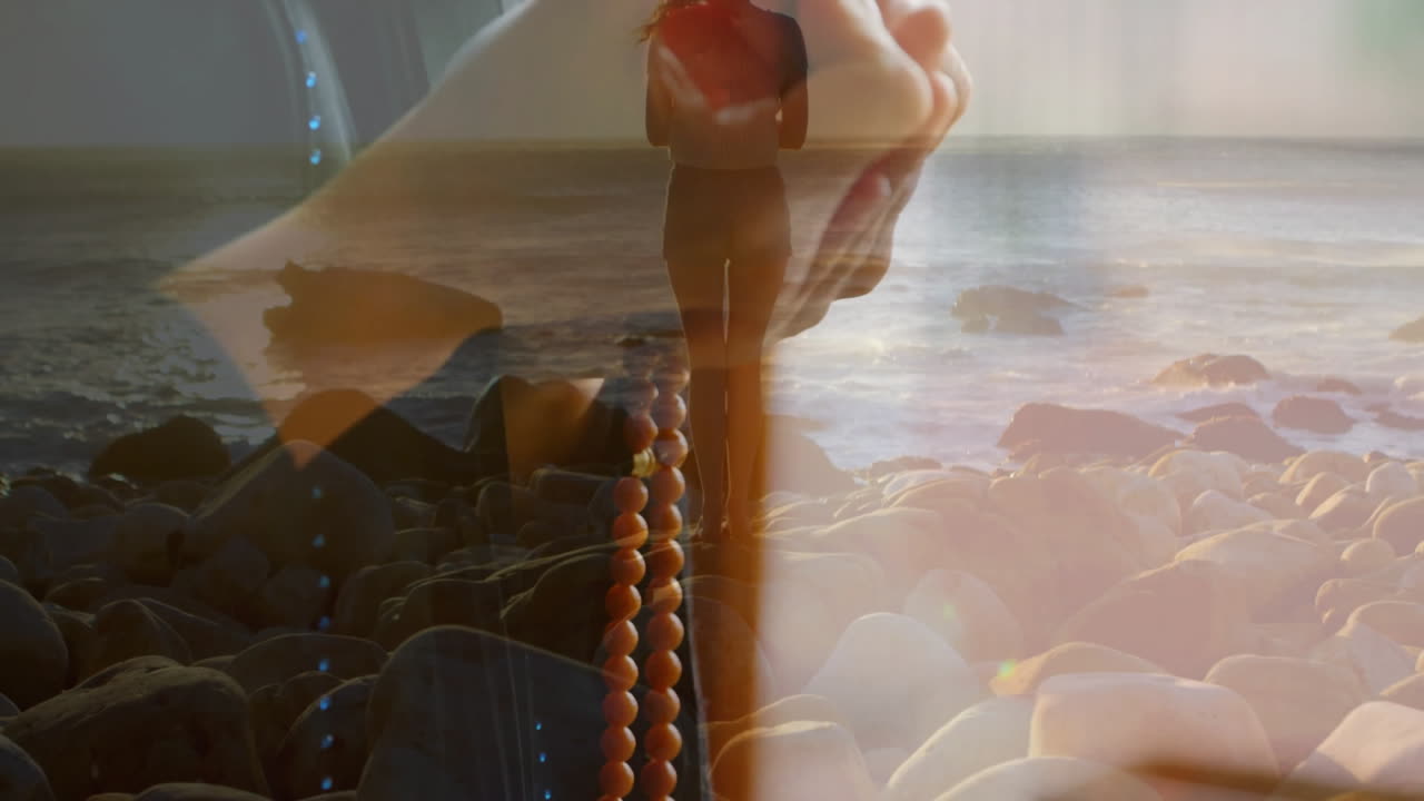 Holding beads, woman standing on rocky beach with ocean waves in background