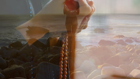 holding beads, woman standing on rocky beach with ocean waves in background