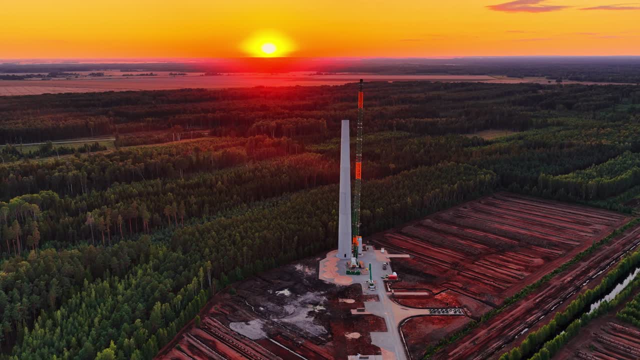 Korkea tuuliturbiinin torni kohoaa vilkkaalta rakennustyömaalta laajan metsä- ja suoalueen keskellä, ja elävä oranssi auringonlasku valaisee maisemaa lämpimällä valolla.