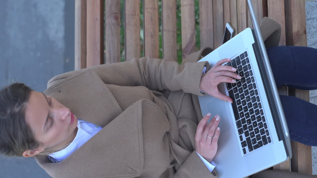 Woman in a brown coat working on her laptop on a bench in the park. Vertical