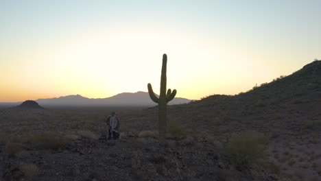 a spaghetti western hero stands on a hill next to a large saguaro cactus and watches the glow of the rising sun illuminate the sonoran desert landscape. the aerial camera orbits the character.