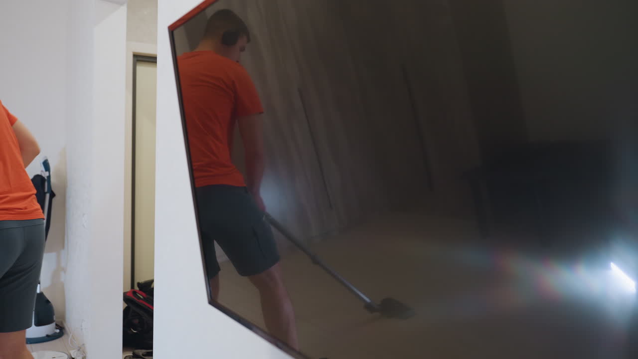 Man in orange shirt and shorts wearing headphones multitasking by dancing and vacuuming wooden floor, reflection visible on TV screen, concept of joyful household cleaning and multitasking