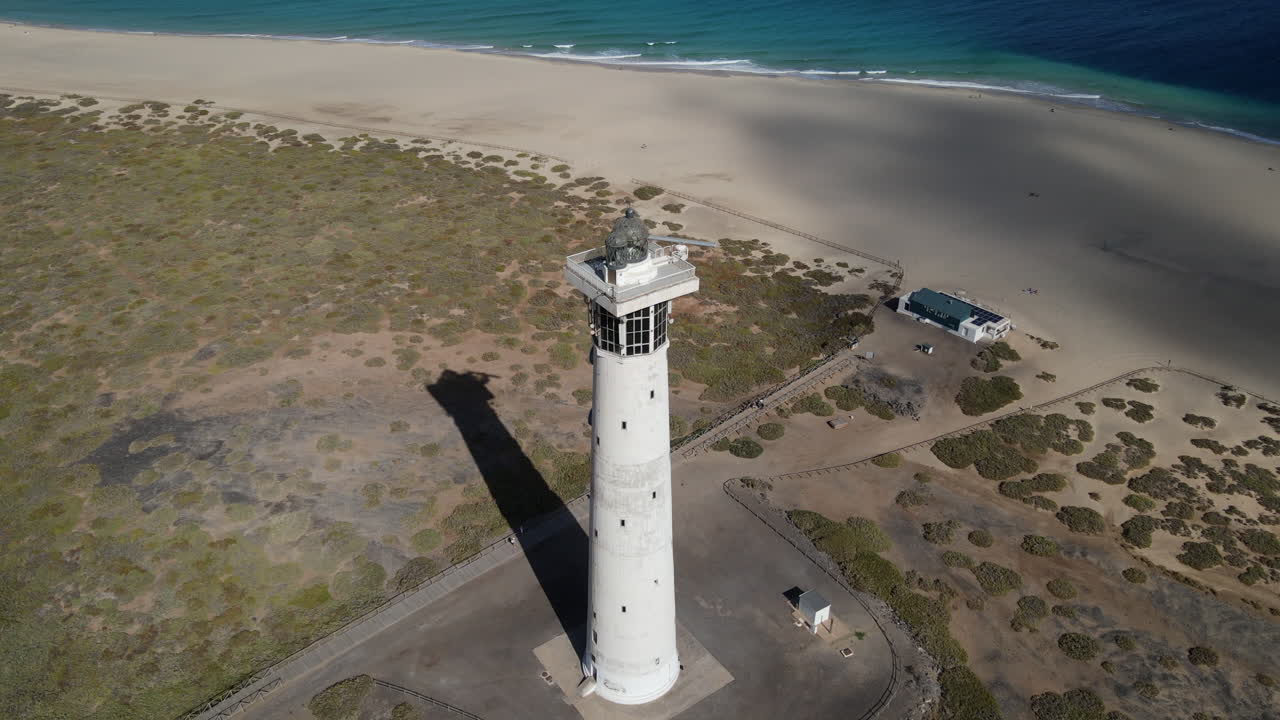 toma aerea en orbita ya media distancia del faro de morro jable, con la playa de morro jable y el mar azul intenso apareciendo al fondo