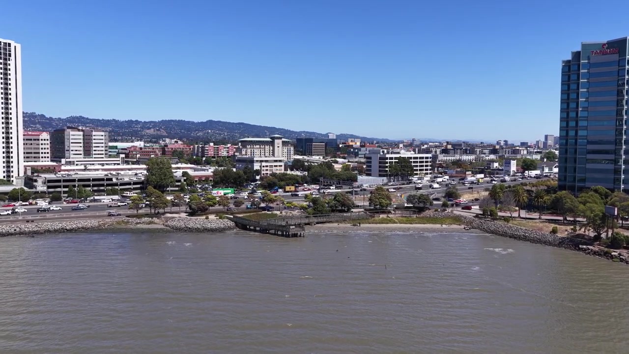 Drone avanza lentamente sopra la baia di Emeryville, CA, tra un grattacielo residenziale e una torre per uffici in vetro, California, stile di vita della Bay Area e sviluppo urbano.