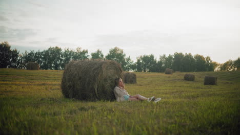 woman seated on ground with legs crossed thoughtfully, resting back against large hay bale in open farmland, peaceful rural landscape with scattered hay bales, warm evening light