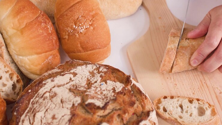 A person slices crusty bread on a wooden board surrounded by various artisan bakery loaves.