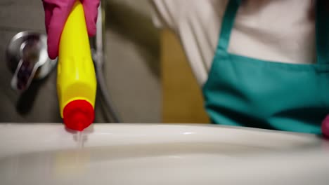 primer plano de una mujer de limpieza confiada en una camiseta blanca y un delantal azul rociando detergente en las paredes del inodoro durante la limpieza en el baño en un apartamento moderno