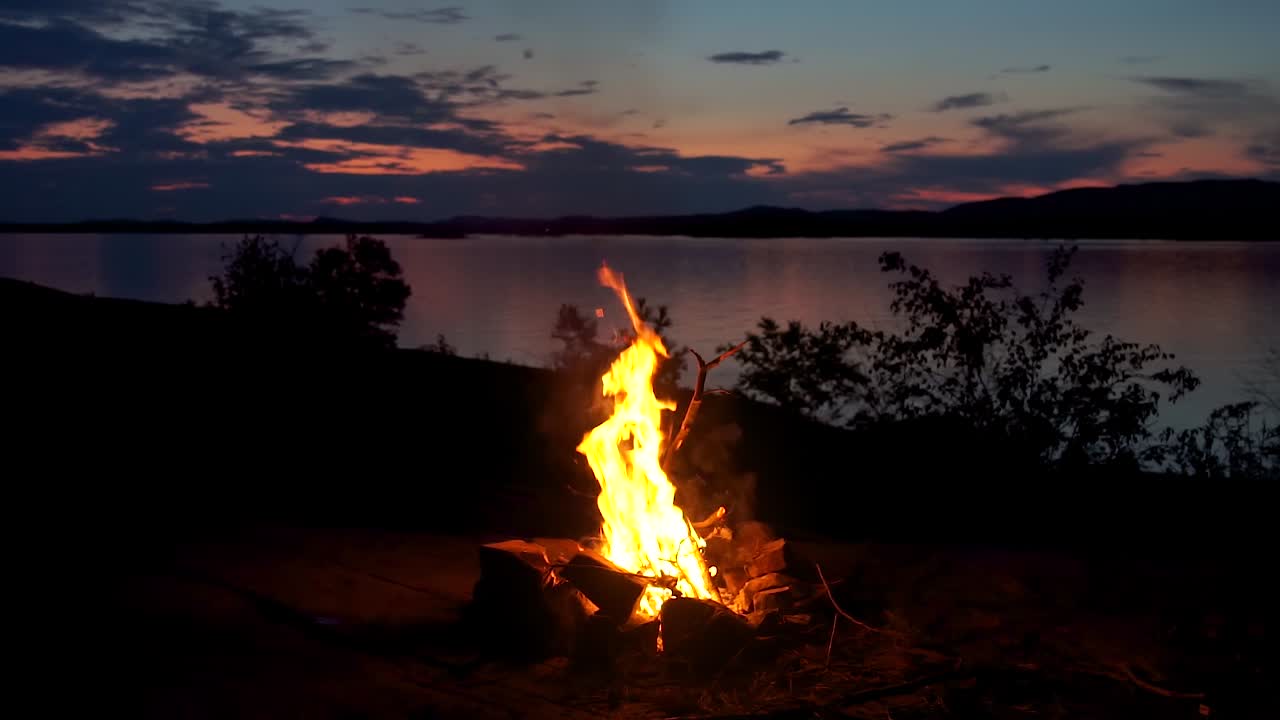 increíble hoguera junto al hermoso lago en canadá en una noche fría con la puesta de sol, las montañas y las nubes en el fondo - toma panorámica