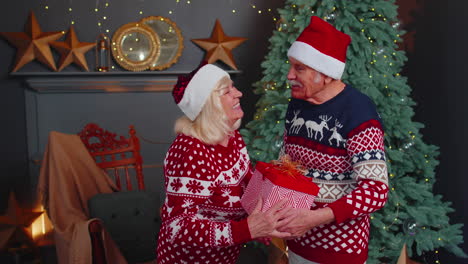 Grandfather-gifting-Christmas-present-box-to-surprised-grandmother-senior-couple-in-Santa-Claus-hats