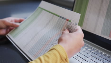 Midsection-of-biracial-woman-sitting-at-desk-using-laptop-and-holding-documents-at-home,-slow-motion