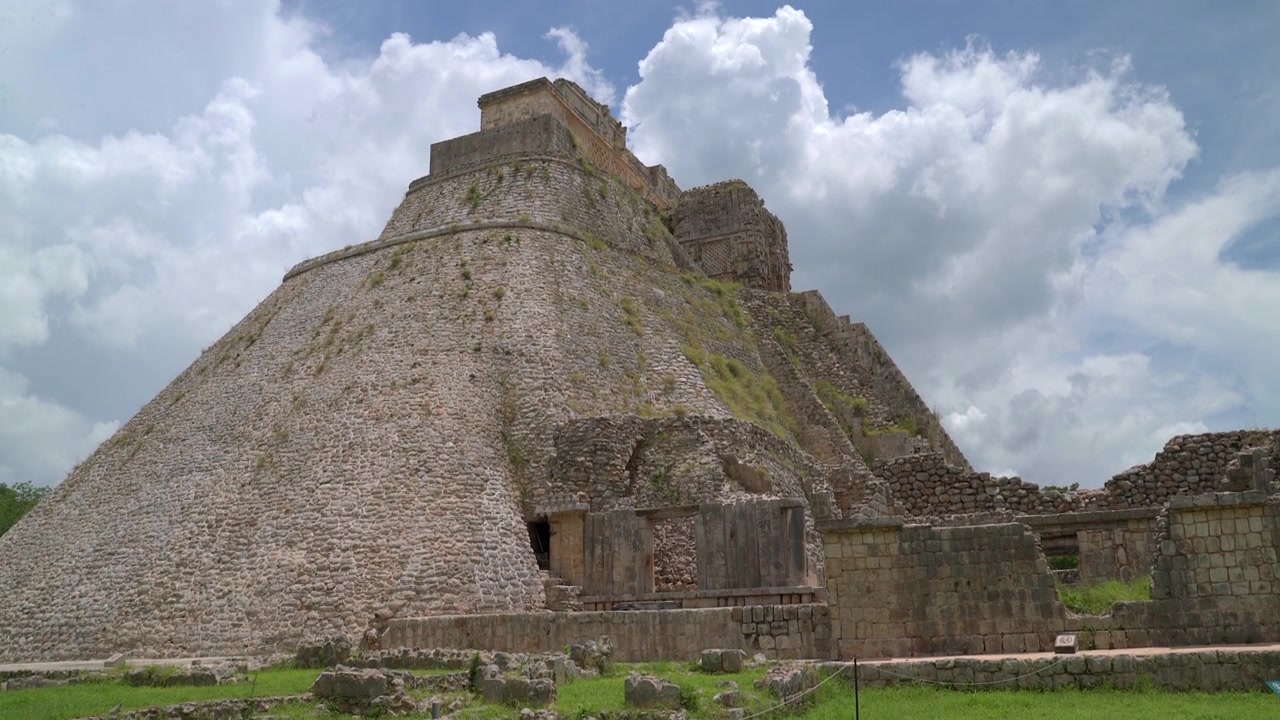 Inquadratura fissa grandangolare dal basso dell'iconica Piramide dell'Indovino di forma ovale presso le rovine Maya di Uxmal nello Yucatan, Messico, sotto un cielo blu nuvoloso.