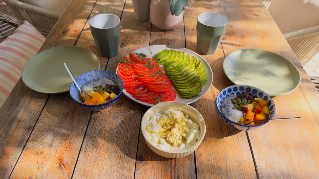 Avocado and tomato slices, cottage cheese and fruit bowls arranged for a healthy breakfast on a rustic wooden table in natural light