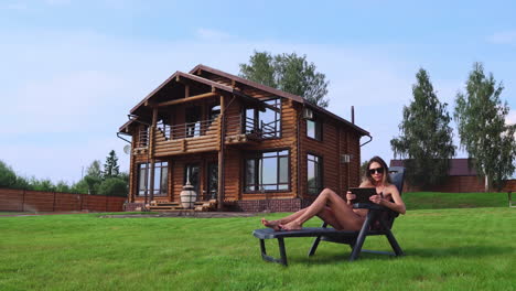 Una-Hermosa-Mujer-Esbelta-Yace-En-Una-Chaise-Longue-En-Traje-De-Baño-Con-Una-Tableta-En-El-Fondo-De-Una-Gran-Casa-De-Campo-Con-Gafas-De-Sol-Y-Toma-El-Sol-Viendo-Fotos-En-Las-Redes-Sociales