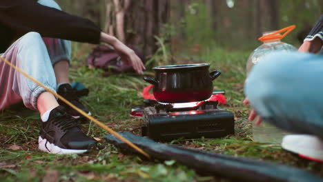 partial view of campers seated in forest adjusting flame on portable gas stove with black pot on top while water bottle sits nearby on ground surrounded by trees, grass, and camping gear
