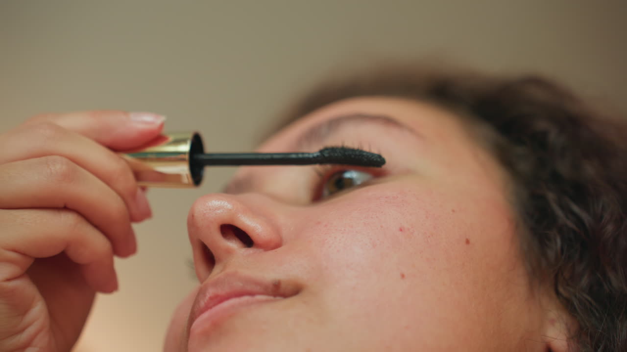 Hotel vanity closeup latina guest mascara applying black mascara on upper lashes, soft warm lighting, serene morning mood, hotel room bed blurred in background, natural skin texture, curly hair, slow