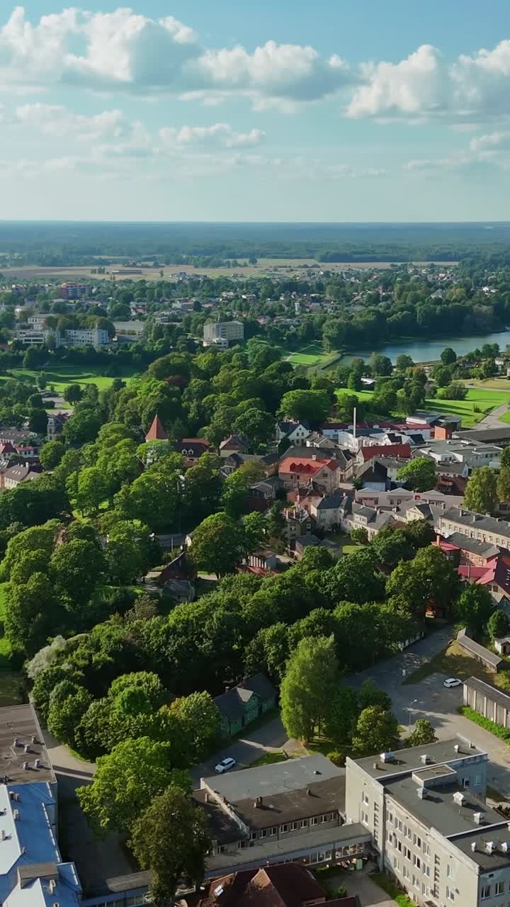 Latvian town Talsi revealing historic roman church, verdant landscape, and winding river from aerial perspective against cloudy background, vertical drone gliding slow