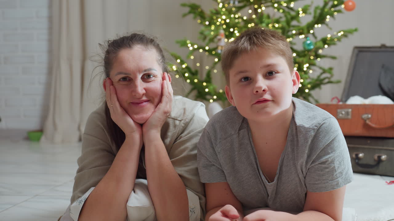 mother and son lying on white rug focusing intently smiling slightly cozy home environment christmas tree with lights in background ornaments scattered nearby festive indoor setting