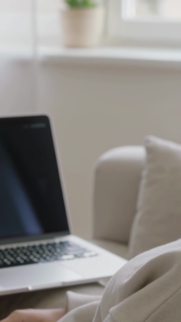 Video verticale: Adulto che si sistema la felpa con cappuccio, seduto sul divano a destra a casa, appoggia la mano vicino a un laptop argentato