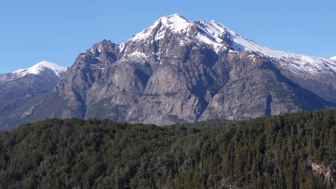 Zoom aereo in allontanamento delle montagne innevate delle Ande con foreste e il Lago Nahuel Huapi vicino a Bariloche, Patagonia.