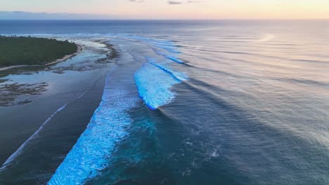 Drone-shot-tilting-up-from-coral-reef-and-island-coastline-with-dense-jungle-and-rolling-waves,-to-the-distant-horizon-at-golden-sunset