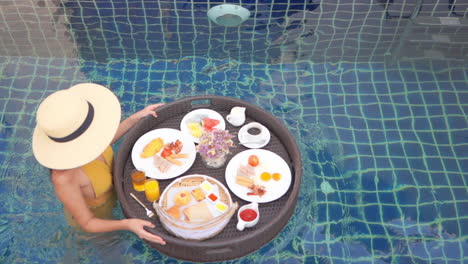 woman in swimsuit walking in pool water with floating plate and served breakfast in luxury hotel resort, slow motion