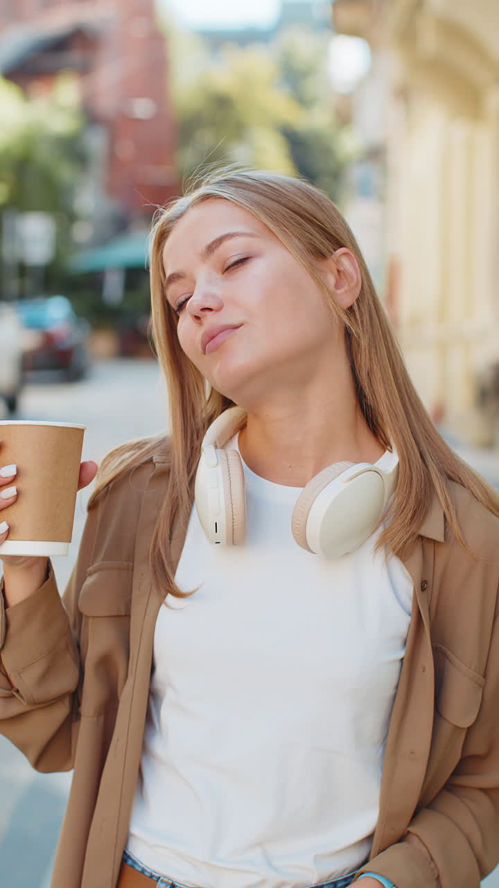 행복한 백인 젊은 여성이 아침 커피를 즐기고 뜨거운 음료를 마시며 웃으며 도시 거리를 고 있습니다.