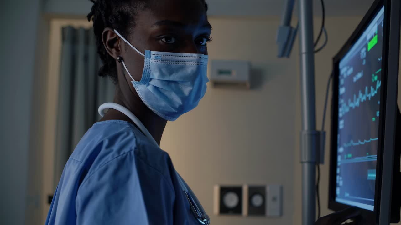 Un operatore sanitario in camice e mascherina controlla un monitor in una stanza poco illuminata.