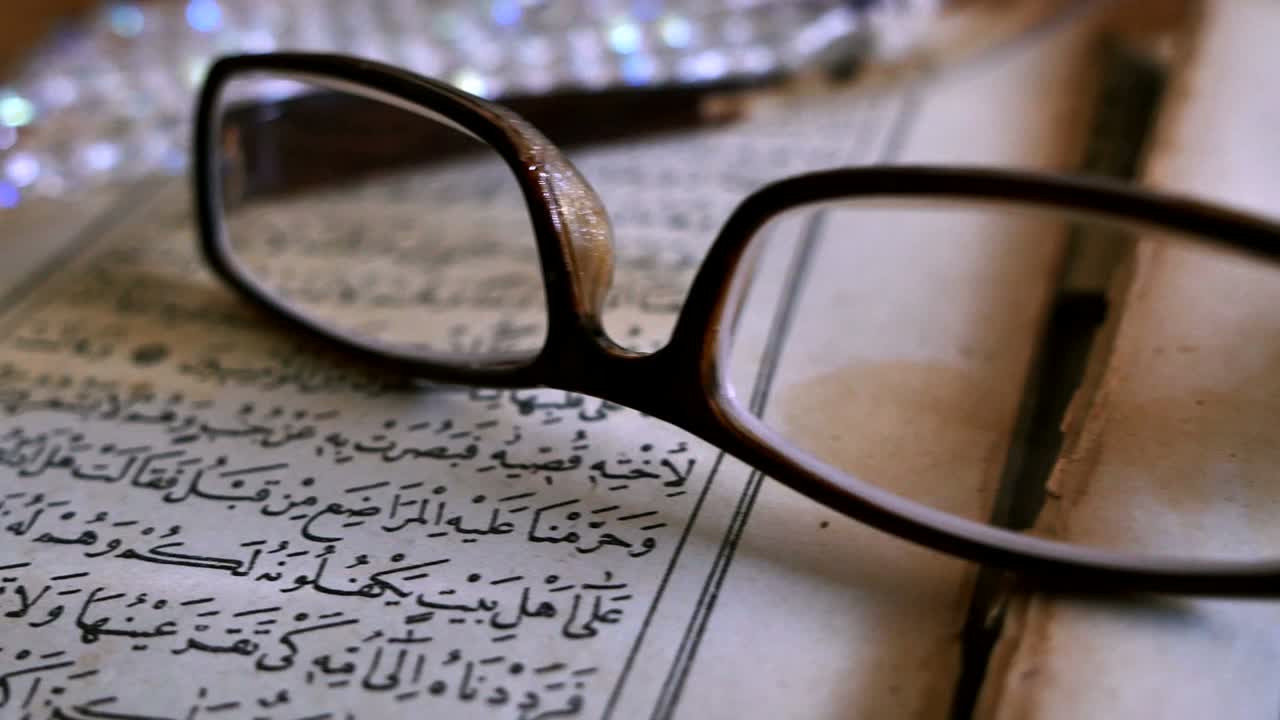 Footage of a Quran lying on a table with reading glasses on it and a tasbeeh in the back, the shot is moving from the right to the left