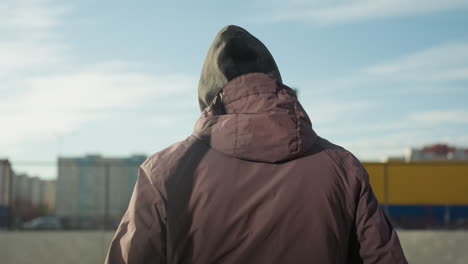 back view of man walking into bar arena holding soccer ball, wearing hoodie and jacket, against blur background of urban residential buildings and vibrant colors, illuminated by bright