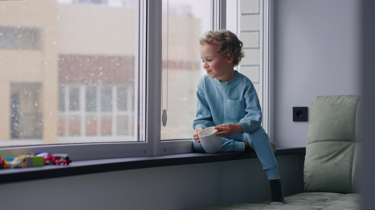 En søt, lubben gutt i blå klær ser på den første snøen i vinduet og drikker melk fra en kopp hjemme.