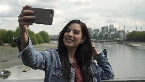 Beautiful-Latina-having-fun-while-taking-a-selfie-with-her-phone,-posing-and-having-a-great-time-while-standing-on-a-bridge-with-a-view-of-London