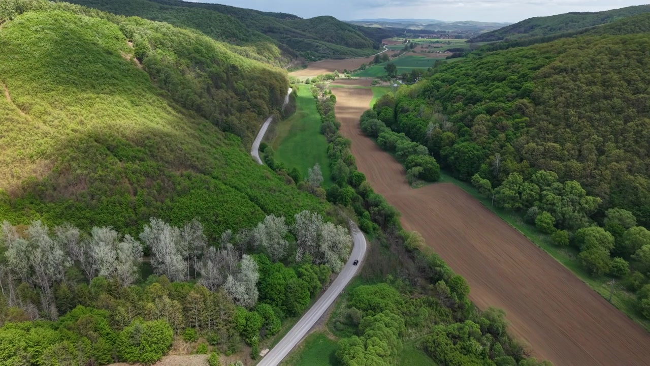 Un'unica auto nera percorre una strada tortuosa circondata da lussureggianti colline verdi, terreni agricoli e fitte foreste primaverili.