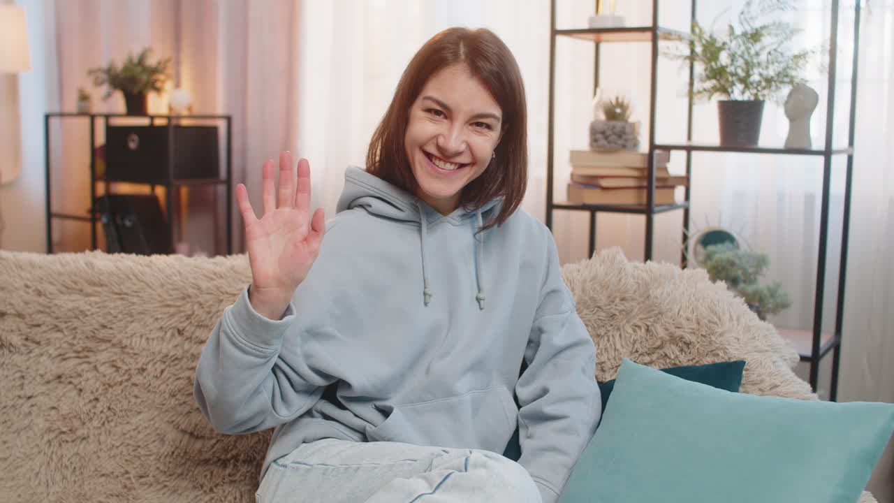 Young caucasian woman girl on sofa joyfully exclaims hey you and waves hand palm in hello gesture