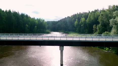 Aerial-Panoramic-Drone-Zoom-out-asphalted-bridge-road-above-river-canal-forest-of-green-pines-and-blue-skyline-around-countryside-transportation-crossing-water-channel