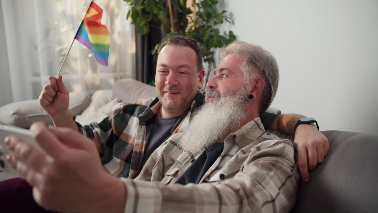 una pareja feliz de hombres lgbt de mediana edad, una morena con una camisa a cuadros y un hombre con cabello gris y una exuberante barba blanca, están sentados en un sofá y tomando una selfie en un apartamento moderno con una bandera lgbt.
