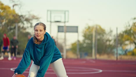 Nahaufnahme-Eines-Blonden-Mädchens-In-Einem-Blauen-Kapuzenpullover-Und-Weißen-Hosen,-Das-Im-Sommer-Einen-Orangefarbenen-Basketball-Auf-Dem-Roten-Boden-Eines-Straßenplatzes-Abprallen-Lässt