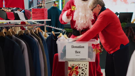 employee dressed as santa claus collecting unneeded clothes from shoppers in donation box to give as gift to unfortunate people during christmas season, spreading holiday cheer