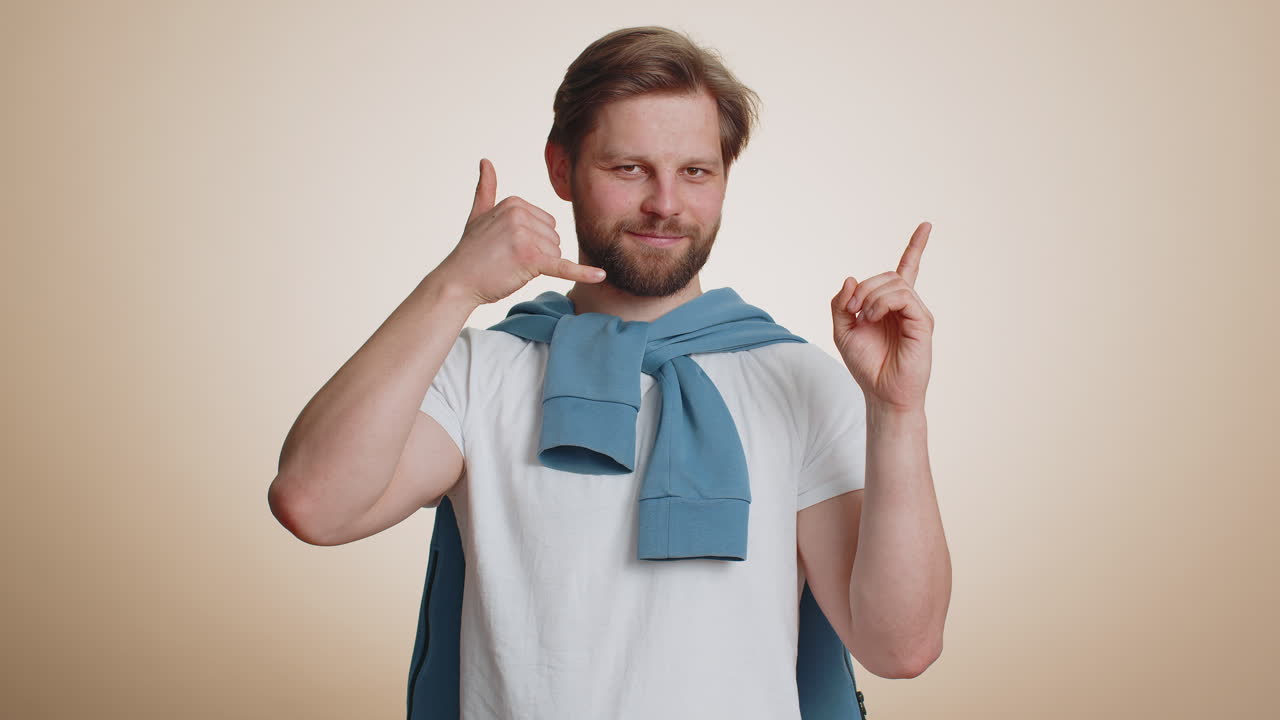 수염이 있는 젊은 남자가 카메라를 바라보며 전화 제스처를 하고 있다.