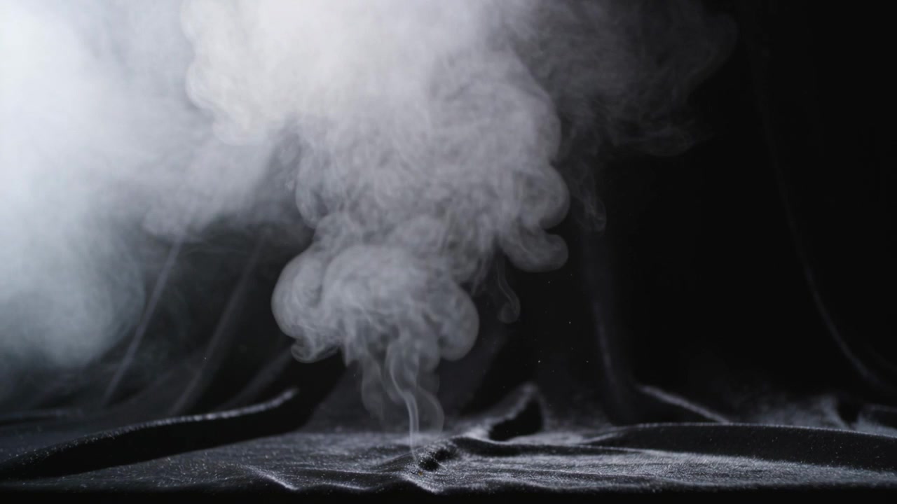 Sorgente nascosta che emette fumo bianco che sale su tessuto scuro drappeggiato in studio.