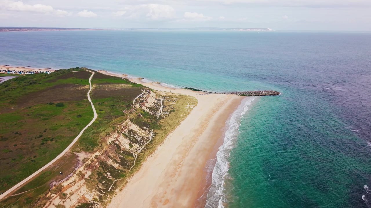 Droneoptagelser over Hengistbury Head med sandstrand, turkisblåt hav, kystklipper, stier, hede, stenmole og den fjerne kystlinje af Isle of Wight.