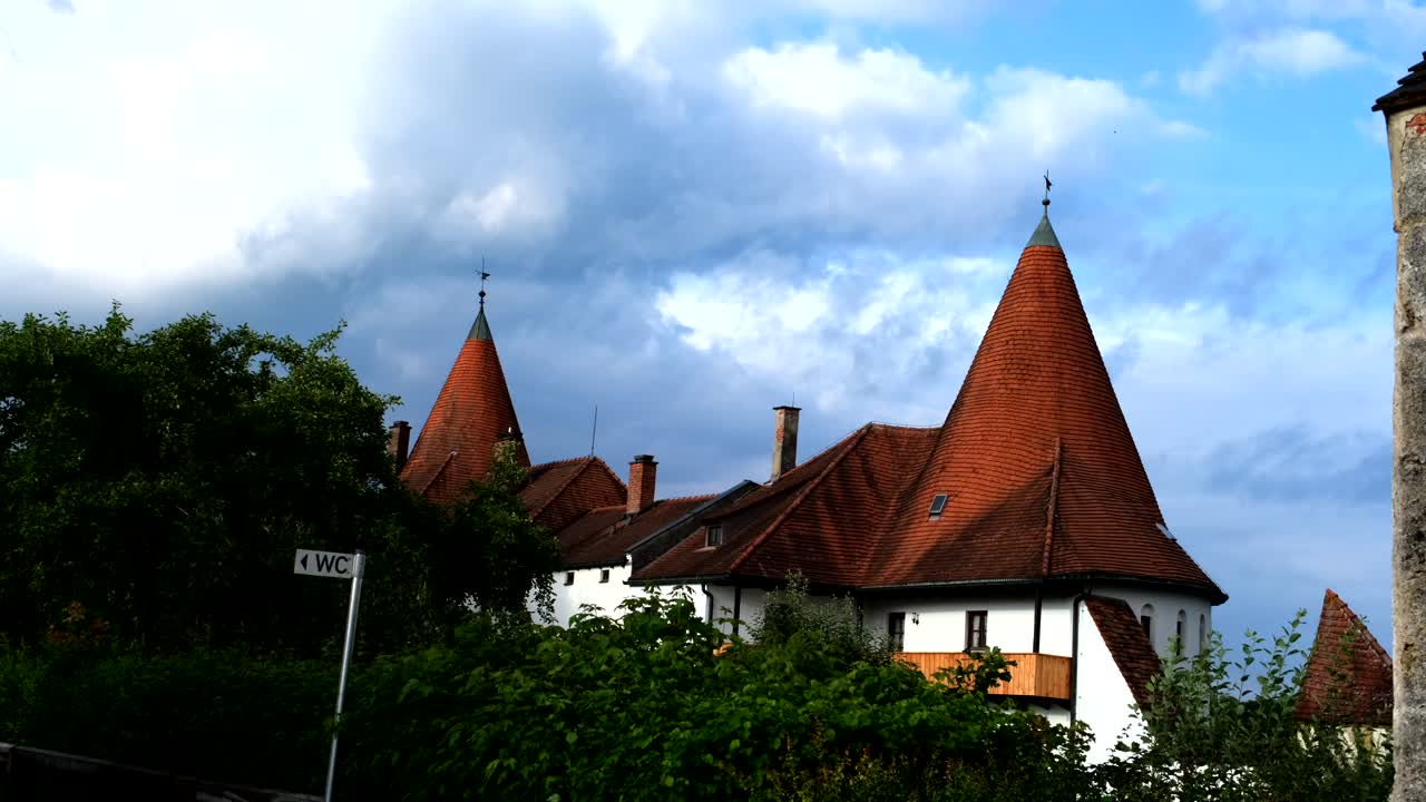 Pointed medieval towers with steep red-tiled roofs rise above lush greenery, set against a backdrop of dramatic clouds and blue sky in a charming historic town