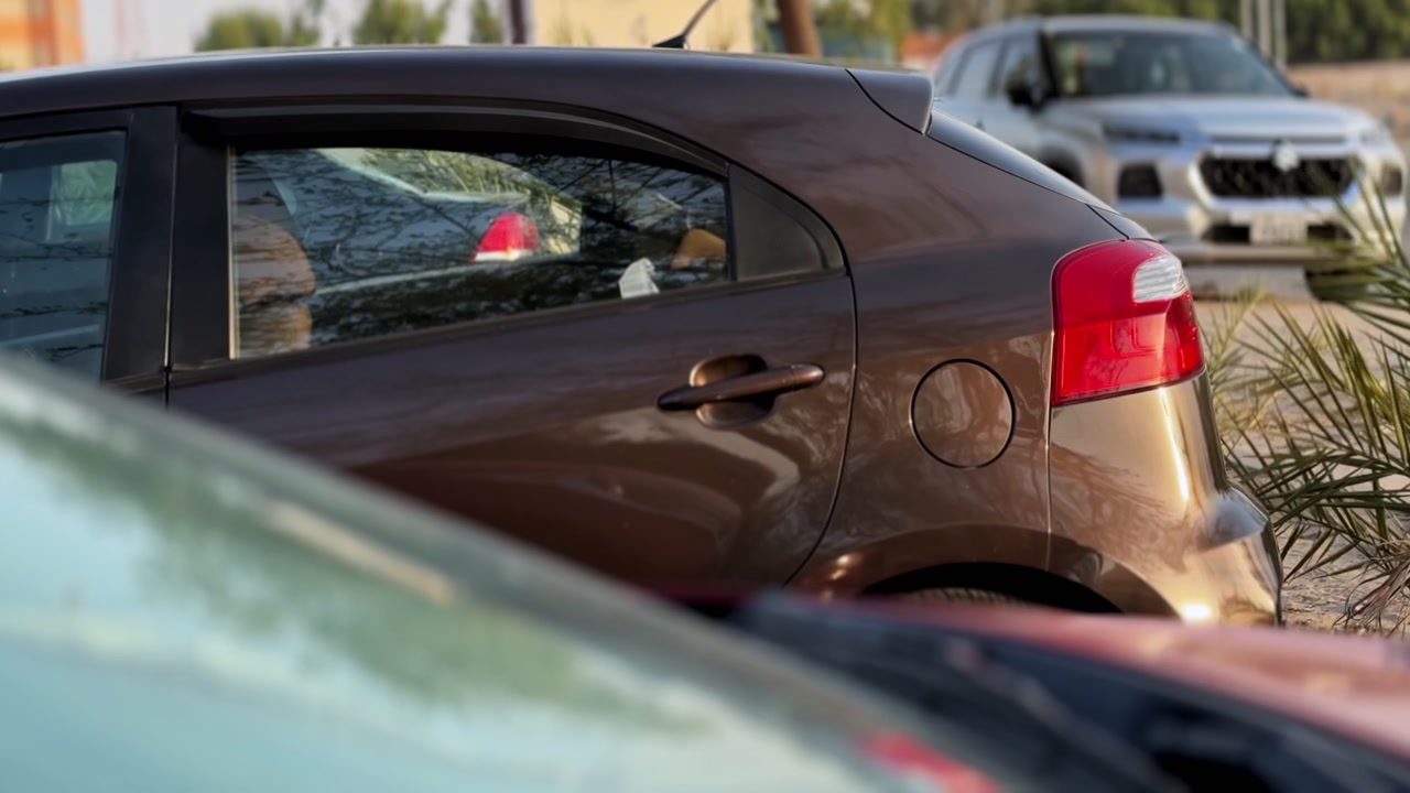Vista lateral de un hatchback compacto marrón estacionado al borde de una carretera bajo la luz cálida del día, con un SUV plateado y otros vehículos suavemente desenfocados al fondo