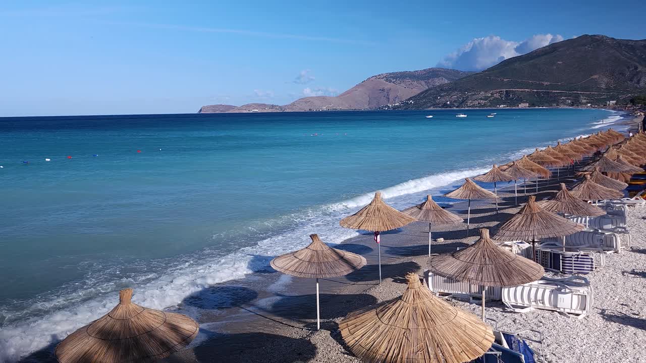 Large beach with straw umbrellas and lounges in Albania, blue sea and white waves on summer holiday panorama