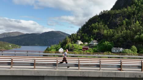 Eye-level-aerial-following-man-jogging-on-Norway-bridge-over-fjord-60-fps